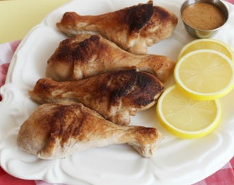 May include: Four baked chicken drumsticks with a browned exterior are arranged on a white, scalloped-edge plate. Slices of yellow lemons and a small metal bowl of sauce are also on the plate. The plate rests on a red and white checkered tablecloth.