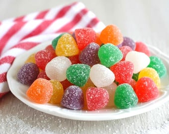 May include: A white plate piled high with assorted gumdrop candies coated in granulated sugar. The candies are dome-shaped and come in various colors including red, orange, yellow, green, white, and purple, creating a vibrant, textured display.