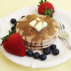 Puede incluir: Una pila de panqueques con sirope y una porción de mantequilla, servidos en un plato blanco con un tenedor. Fresas y arándanos frescos adornan los panqueques, creando una escena de desayuno colorida y apetitosa.