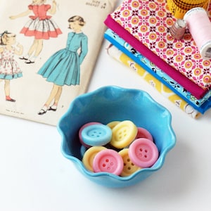 May include: A blue ceramic bowl filled with colourful buttons. The buttons are in shades of pink, blue, and yellow. The bowl is sitting on a white surface.