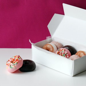 Puede incluir: Una caja blanca llena de donuts variados, algunos con chispas. Dos donuts están fuera de la caja. Uno es rosa con chispas coloridas, y el otro es de chocolate. El fondo es de color magenta.