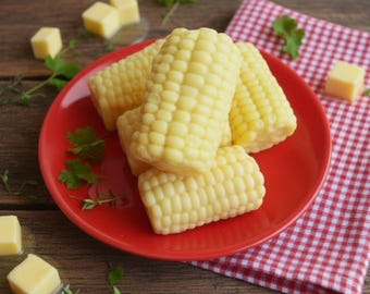 May include: A red plate holds several cooked corn on the cob, with yellow kernels. Cubes of butter and sprigs of herbs are scattered around the plate. A red and white checkered napkin is in the background.
