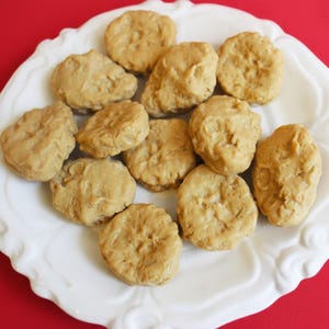 Könnte beinhalten: Ein weißer Teller mit gewelltem Rand, gefüllt mit einem Dutzend goldbraunen, unregelmäßig geformten Lebensmitteln. Das Essen hat eine strukturierte Oberfläche und scheint gebraten oder gebacken zu sein. Der Teller steht auf rotem Untergrund.