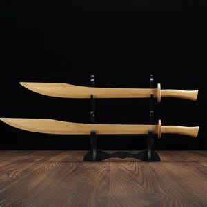 May include: Two wooden swords with curved blades and rounded handles are displayed on a black stand. The swords are light brown, and the stand is black. The background is a dark brown wooden surface and a black backdrop.