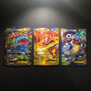 Puede incluir: Tres cartas coleccionables Pokémon EX con Venusaur, Charizard y Blastoise. La carta de Venusaur tiene una combinación de colores verde y azul, la carta de Charizard es naranja y amarilla, y la carta de Blastoise es azul y blanca. Cada carta tiene un fondo holográfico.