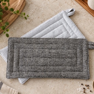 May include: Two rectangular pot holders featuring a quilted texture and a fabric loop for hanging. One pot holder is a dark charcoal grey with a woven appearance, while the second one underneath is a lighter heather grey fabric.