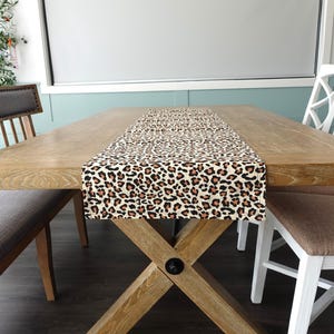 May include: A wooden dining table with a leopard print table runner. The table has a cross-shaped base and is set with two chairs.