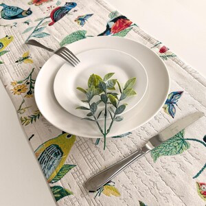 May include: A white table runner with a floral pattern featuring birds and butterflies. Two white plates are stacked on top of each other with a small sprig of green leaves in the centre. A silver knife is placed on the table runner.