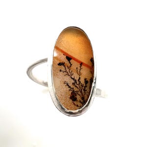 Puede incluir: Anillo de plata con una piedra preciosa ovalada que muestra un patr&oacute;n natural en forma de &aacute;rbol en tonos marr&oacute;n, naranja y negro. El anillo tiene un dise&ntilde;o simple y elegante, que resalta la belleza &uacute;nica de la piedra.