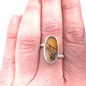 Puede incluir: Un anillo de plata con una piedra ovalada que muestra un patr&oacute;n natural en tonos beige, marr&oacute;n y negro. La piedra est&aacute; engastada en una sencilla banda de plata, creando un dise&ntilde;o minimalista. El anillo se muestra en una mano.