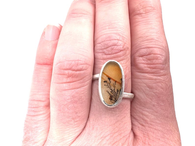 Puede incluir: Un anillo de plata con una piedra ovalada que muestra un patr&oacute;n natural en tonos beige, marr&oacute;n y negro. La piedra est&aacute; engastada en una sencilla banda de plata, creando un dise&ntilde;o minimalista. El anillo se muestra en una mano.