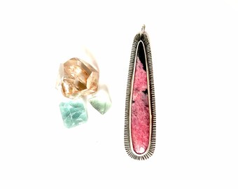 May include: A silver pendant with a pink and black stone set in a silver frame. The pendant is hanging from a silver chain.