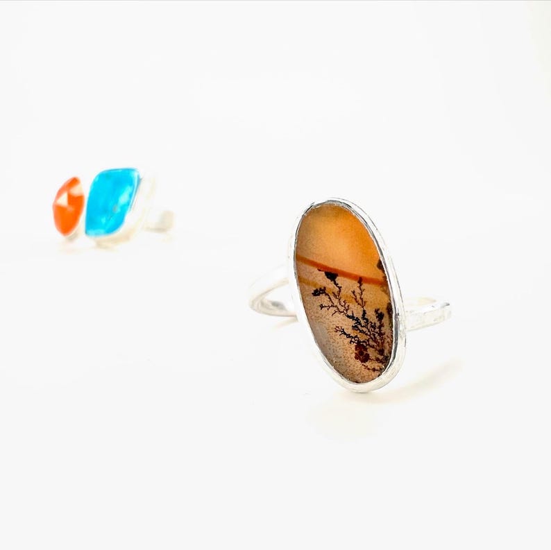 Puede incluir: Un anillo de plata con una piedra ovalada de color &aacute;mbar con un patr&oacute;n negro en forma de &aacute;rbol. En el fondo se ve otro anillo con una piedra turquesa y naranja. Los anillos est&aacute;n sobre un fondo blanco.