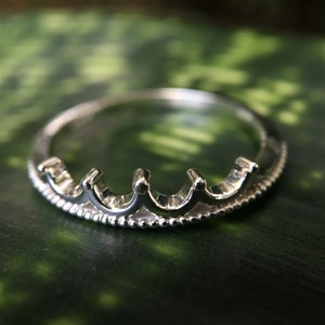 Peut inclure: Bague argent&eacute;e en forme de couronne. La bague pr&eacute;sente une s&eacute;rie de petites arches arrondies sur le dessus, avec une texture perl&eacute;e sur le bord inf&eacute;rieur. La bague est pos&eacute;e sur un fond vert.