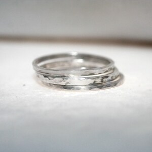 May include: Four silver rings with a hammered finish are stacked together. The rings have a simple band design with a textured surface. The rings are made of silver and have a handcrafted appearance. The rings are stacked on top of each other.