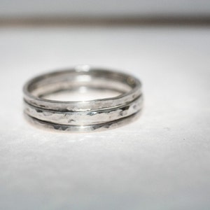 May include: Three silver rings with a hammered finish, stacked together. Each ring is a simple band with a textured surface, catching the light. The rings are stacked, creating a layered effect. The rings are approximately 0.2 cm wide.