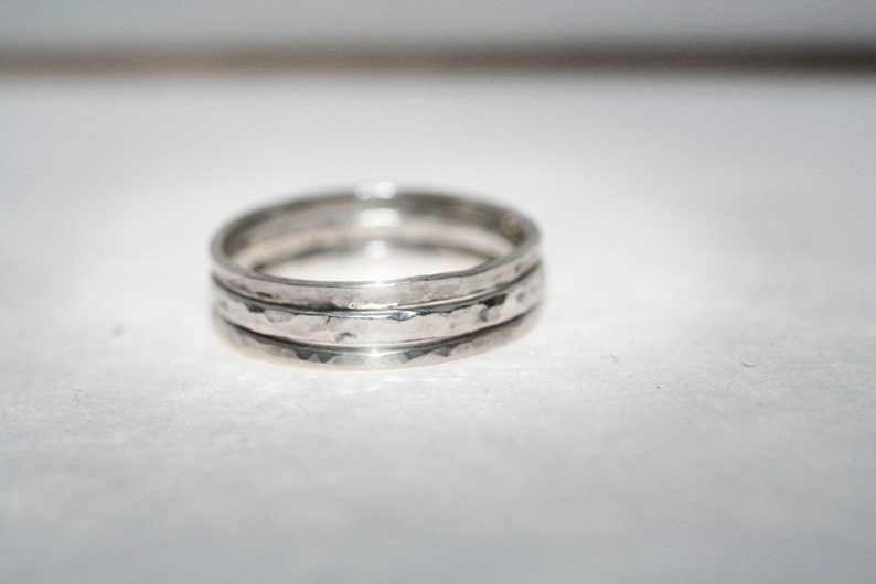 May include: Three silver rings with a hammered finish, stacked together. Each ring is a simple band with a textured surface, catching the light. The rings are stacked, creating a layered effect. The rings are approximately 0.2 cm wide.