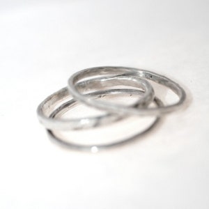 May include: Three silver rings interlocked, creating a unique design. The rings have a textured surface and a slightly irregular shape, giving them a handcrafted appearance. The rings are set against a white background.