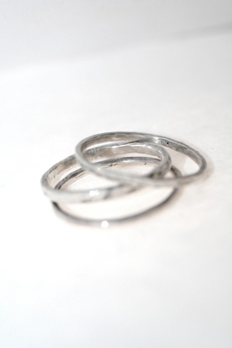 May include: Three silver rings interlocked, creating a unique design. The rings have a textured surface and a slightly irregular shape, giving them a handcrafted appearance. The rings are set against a white background.