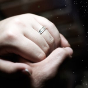May include: A silver ring with a textured design is worn on a person's finger. The ring is the focal point, set against a blurred background of hands and a dark, starry effect.