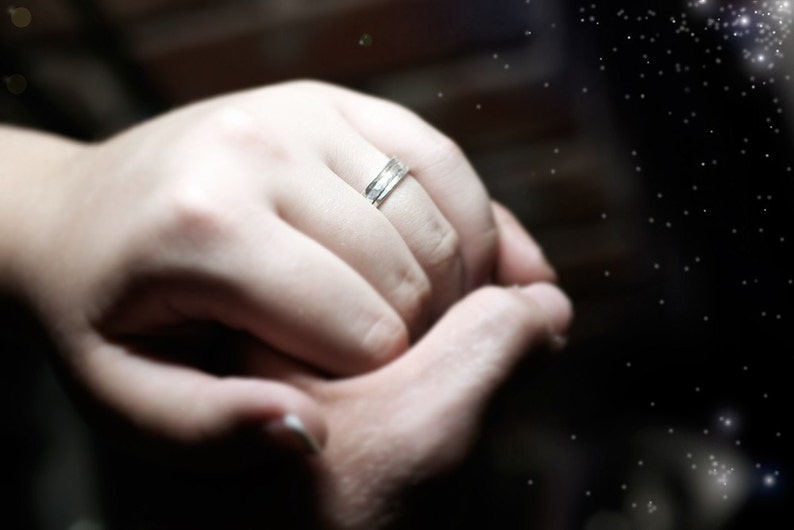 May include: A silver ring with a textured design is worn on a person's finger. The ring is the focal point, set against a blurred background of hands and a dark, starry effect.