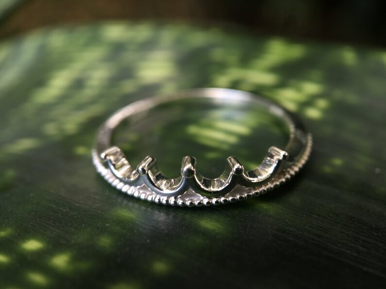 Peut inclure: Bague argent&eacute;e en forme de couronne. La bague pr&eacute;sente une s&eacute;rie de petites arches arrondies sur le dessus, avec une texture perl&eacute;e sur le bord inf&eacute;rieur. La bague est pos&eacute;e sur un fond vert.