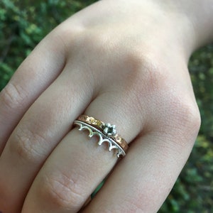 Peut inclure: Ensemble de deux bagues. La bague inf&eacute;rieure est argent&eacute;e avec un motif de couronne. La bague sup&eacute;rieure est dor&eacute;e avec une texture martel&eacute;e. Une petite gemme argent&eacute;e est pos&eacute;e sur la bague dor&eacute;e. Les bagues sont sur le doigt d'une personne.