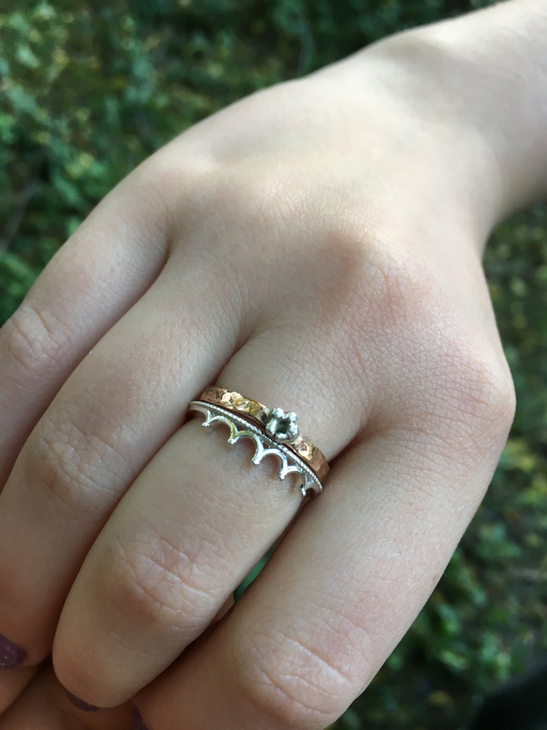 Peut inclure: Ensemble de deux bagues. La bague inf&eacute;rieure est argent&eacute;e avec un motif de couronne. La bague sup&eacute;rieure est dor&eacute;e avec une texture martel&eacute;e. Une petite gemme argent&eacute;e est pos&eacute;e sur la bague dor&eacute;e. Les bagues sont sur le doigt d'une personne.