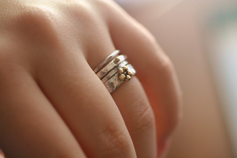 May include: A collection of silver rings with textured surfaces and small gold embellishments. The rings are worn on a finger, highlighting the contrast between the silver bands and the gold details. The rings are a piece of jewellery.