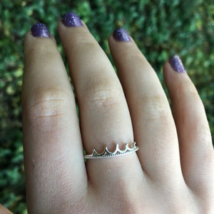 Peut inclure: Une bague couronne argent&eacute;e port&eacute;e au doigt. La bague pr&eacute;sente un motif de couronne d&eacute;licat avec cinq pics. Les ongles de la personne sont peints avec du vernis &agrave; ongles paillet&eacute; violet.