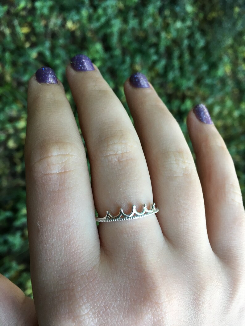 Peut inclure: Une bague couronne argent&eacute;e port&eacute;e au doigt. La bague pr&eacute;sente un motif de couronne d&eacute;licat avec cinq pics. Les ongles de la personne sont peints avec du vernis &agrave; ongles paillet&eacute; violet.