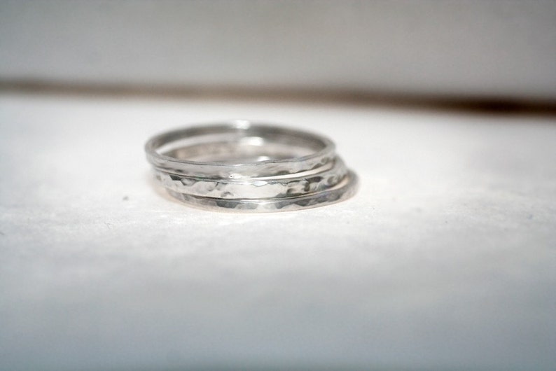 May include: Four silver rings with a hammered finish are stacked together. The rings have a simple band design with a textured surface. The rings are made of silver and have a handcrafted appearance. The rings are stacked on top of each other.
