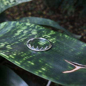 Peut inclure: Une bague en argent avec un motif de couronne. La bague pr&eacute;sente une s&eacute;rie de petits arcs incurv&eacute;s sur le dessus, avec une bordure perl&eacute;e. La bague est pos&eacute;e sur un fond de feuille verte.