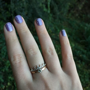 Peut inclure: Une main portant deux bagues, l'une avec un motif de couronne et l'autre avec une petite pierre pr&eacute;cieuse. Les ongles sont peints avec un vernis &agrave; ongles violet scintillant. L'arri&egrave;re-plan est vert flou.