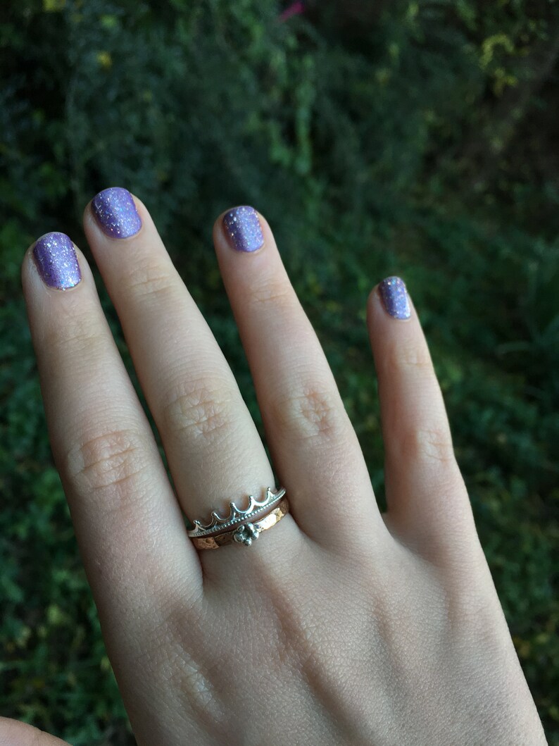 Peut inclure: Une main portant deux bagues, l'une avec un motif de couronne et l'autre avec une petite pierre pr&eacute;cieuse. Les ongles sont peints avec un vernis &agrave; ongles violet scintillant. L'arri&egrave;re-plan est vert flou.