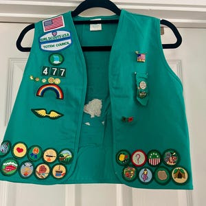 May include: A green Girl Scouts vest with various patches and pins. Patches include the American flag, the words "Girl Scouts USA" and "Totem Council", a rainbow, and several merit badges. The vest is hanging on a black hanger.