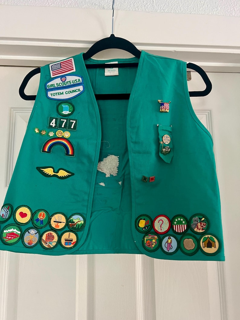 May include: A green Girl Scouts vest with various patches and pins. Patches include the American flag, the words "Girl Scouts USA" and "Totem Council", a rainbow, and several merit badges. The vest is hanging on a black hanger.