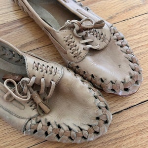 May include: A pair of tan leather shoes with woven detailing and laces. The shoes have a tassel accent on the laces.