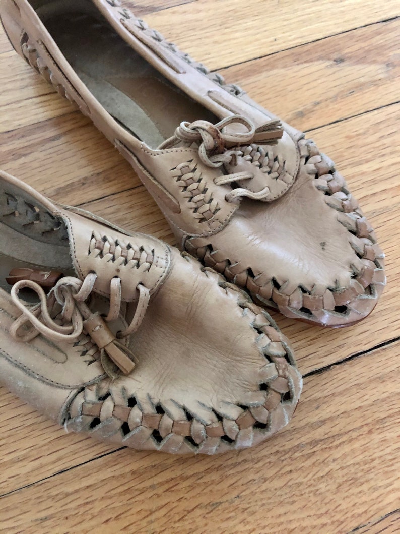 May include: A pair of tan leather shoes with woven detailing and laces. The shoes have a tassel accent on the laces.