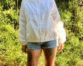 May include: White zip-up jacket with a stand-up collar. The jacket has a zipper closure and two pockets. The model is wearing the jacket with denim shorts.