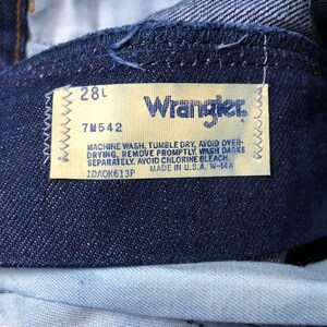 May include: A close-up of a blue denim Wrangler jeans label with white stitching. The label has the size "28L" and the style number "7M542". The label also includes care instructions: "Machine wash, tumble dry, avoid over-drying, remove promptly. Wash darks separately. Avoid chlorine bleach." The label also states "Made in U.S.A. W-14A" and the number "1DA0K613P".