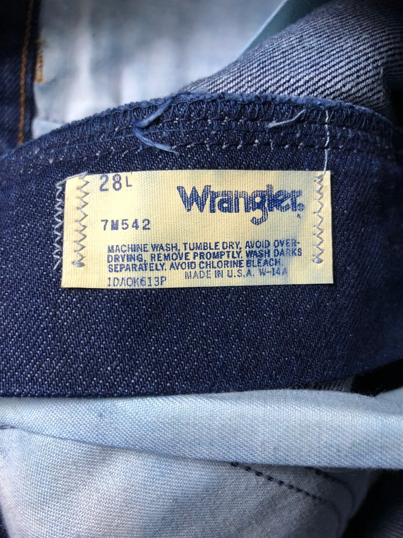 May include: A close-up of a blue denim Wrangler jeans label with white stitching. The label has the size "28L" and the style number "7M542". The label also includes care instructions: "Machine wash, tumble dry, avoid over-drying, remove promptly. Wash darks separately. Avoid chlorine bleach." The label also states "Made in U.S.A. W-14A" and the number "1DA0K613P".