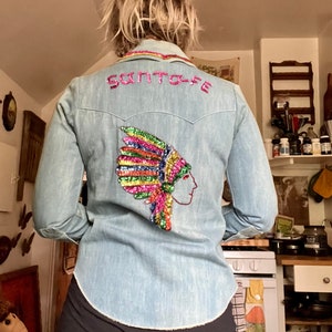 May include: A light blue denim shirt with a sequined Native American headdress design on the back. The shirt has the word "Santa Fe" embroidered on the back in pink sequins.