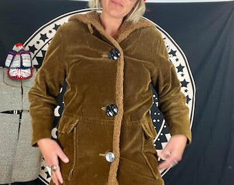 May include: A brown corduroy coat with a faux fur lining and large black buttons. The coat has two front pockets and a hood. The person in the image is wearing a red beanie with a white label.