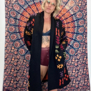 May include: A black knitted cardigan with a colorful geometric pattern. The cardigan has a shawl collar and long sleeves. The model is wearing the cardigan open, revealing a black bra underneath.