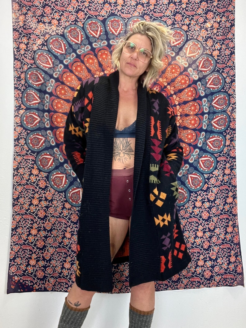 May include: A black knitted cardigan with a colorful geometric pattern. The cardigan has a shawl collar and long sleeves. The model is wearing the cardigan open, revealing a black bra underneath.