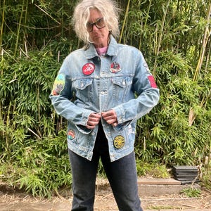 Puede incluir: Chaqueta vaquera azul claro adornada con parches coloridos. La chaqueta presenta un diseño clásico con botones y dos bolsillos en el pecho. Los parches muestran varios diseños. Se usa con vaqueros negros, creando un look personalizado.