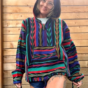 May include: A colourful striped hooded sweatshirt with a geometric pattern. The sweatshirt has a drawstring hood and a kangaroo pocket. The person wearing the sweatshirt is wearing a blue beanie.