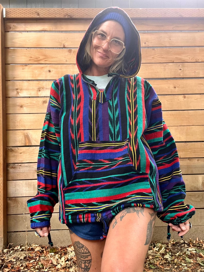 May include: A colourful striped hooded sweatshirt with a geometric pattern. The sweatshirt has a drawstring hood and a kangaroo pocket. The person wearing the sweatshirt is wearing a blue beanie.