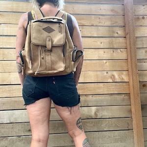 May include: A person wearing denim shorts and a brown leather backpack with a diamond-shaped logo. The backpack has a drawstring closure and multiple pockets. The person has tattoos on their legs.